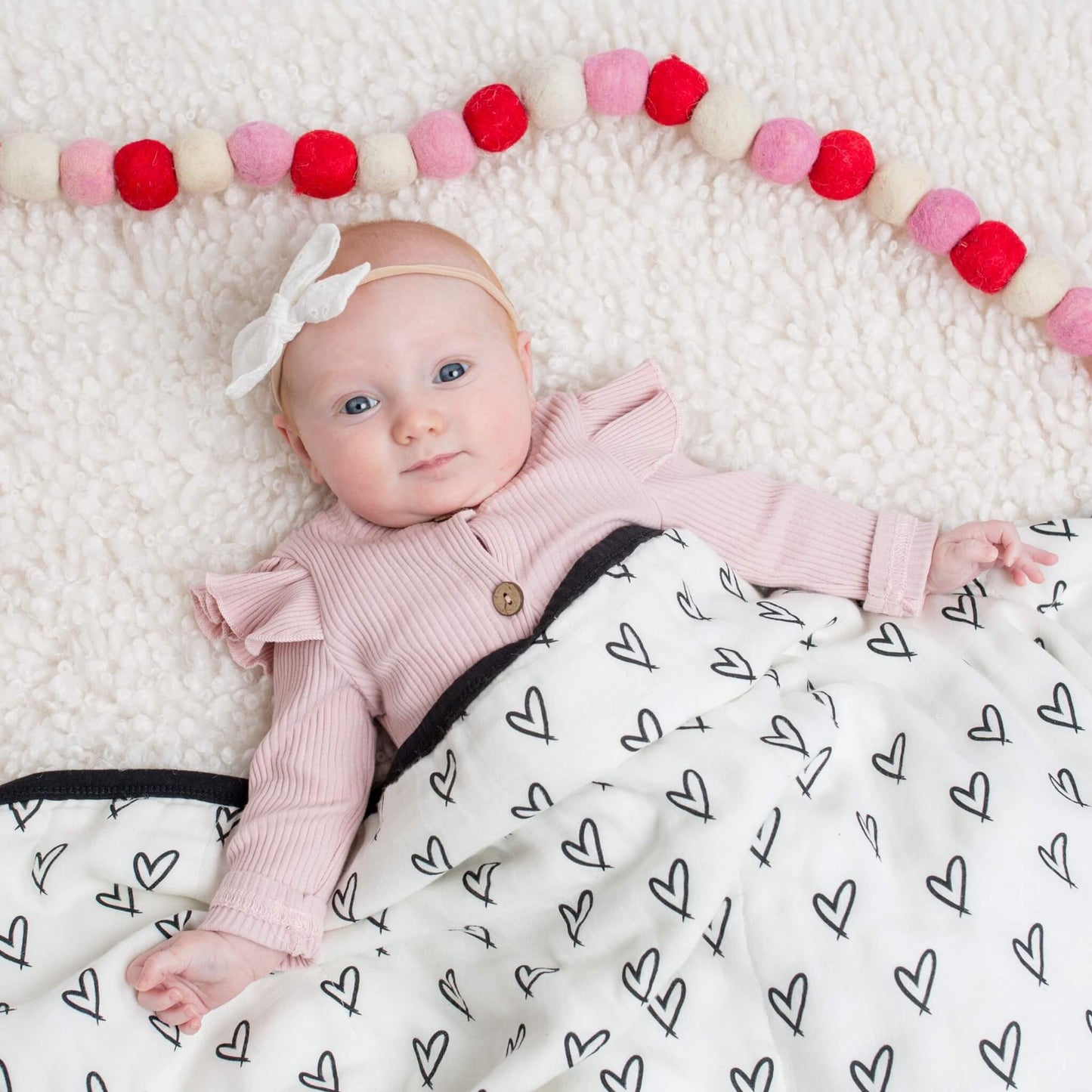 A baby lies on a soft surface, wrapped in the Lulujo Quilted Blanket in Messy Heart print, adding warmth and style.