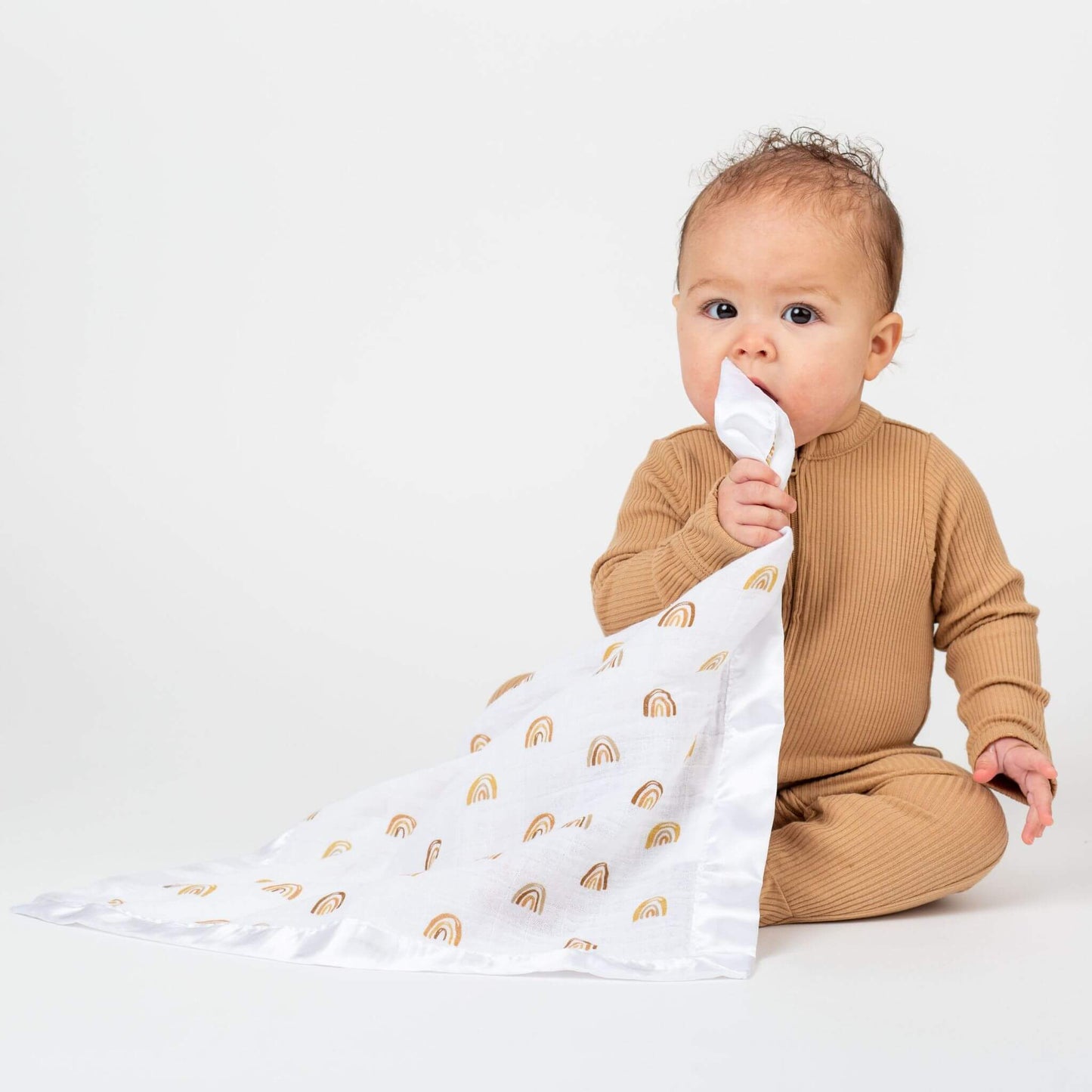 Baby in ribbed caramel outfit mouthing Lulujo Rainbow security blanket with gold pattern on white fabric, demonstrating the blanket's soothing properties.