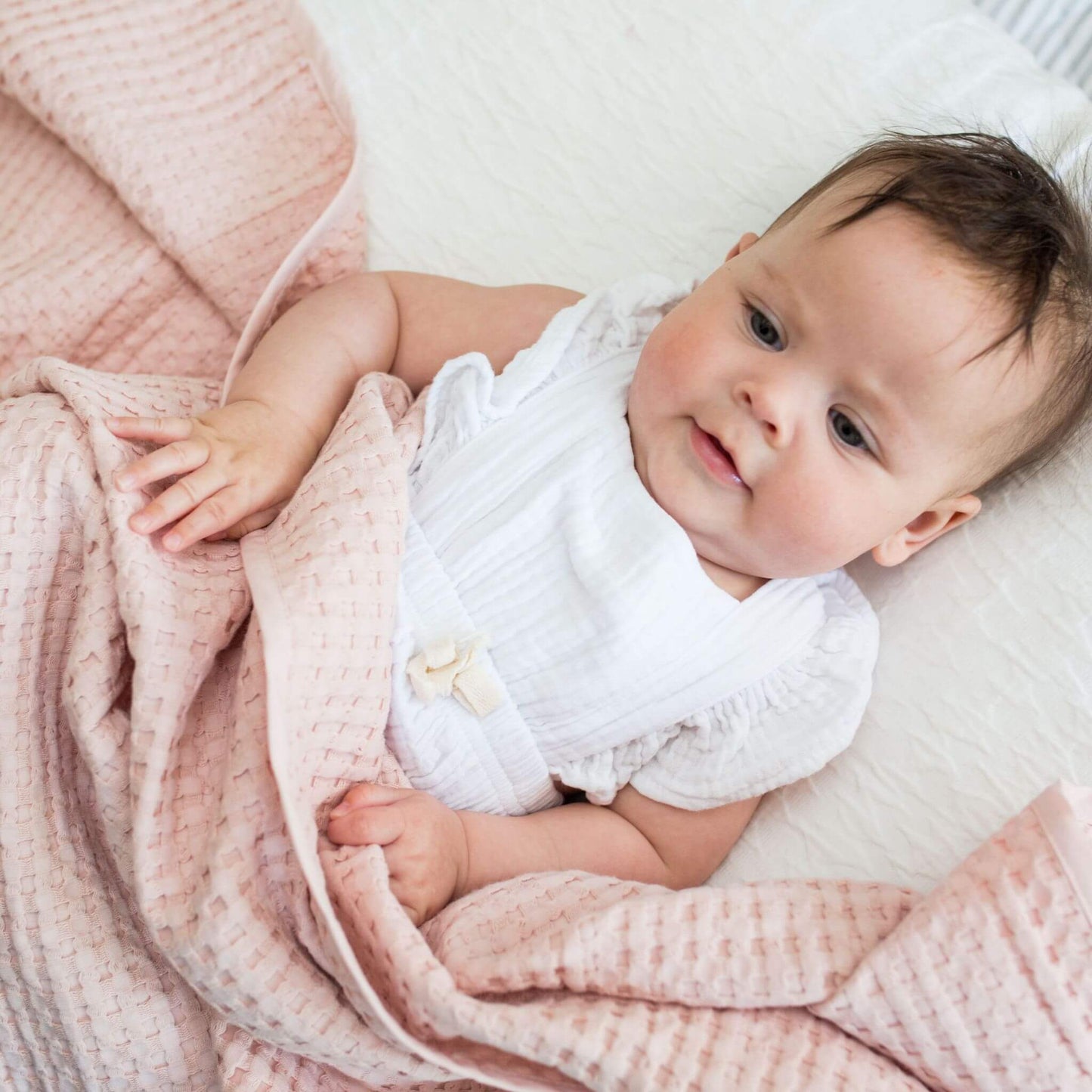 Smiling baby with dark hair in white clothing partially covered with a pink Lulujo waffle blanket while lying on a white surface.