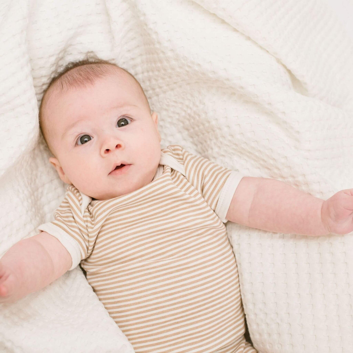 Newborn baby with dark eyes in striped bodysuit lying comfortably on white waffle-textured blanket, arms outstretched and looking upward with an attentive expression.