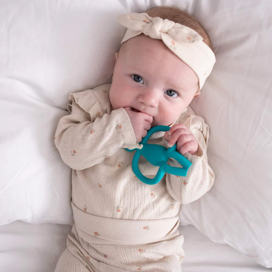A baby lying on a white bed holding a turquoise elephant-shaped teether, chewing on the soft silicone trunk while gripping the round side handles.