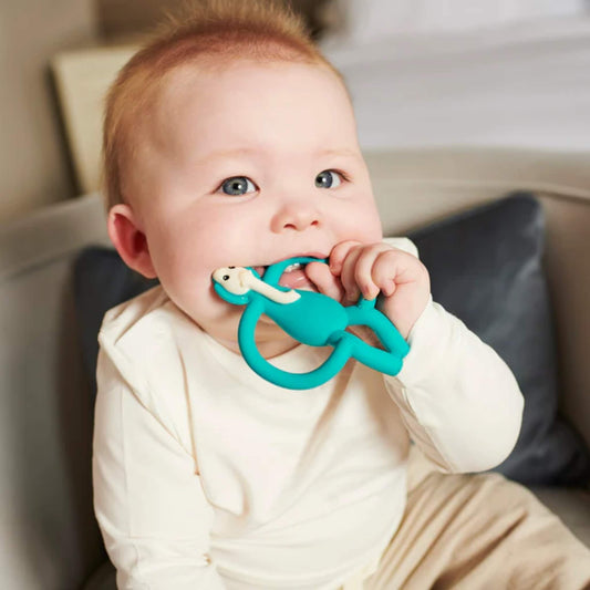 Baby sitting on sofa holding and chewing turquoise elephant silicone teether, demonstrating easy grip handles and soft chewable texture for teething relief.