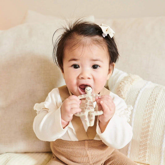 A baby sitting on a soft cushion holding a beige giraffe-shaped silicone teether with both hands while chewing the top section.