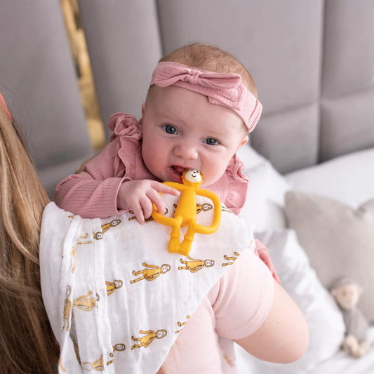 A baby chewing on a yellow lion-shaped silicone teether while being held over a shoulder, with a white muslin patterned with small lion illustrations draped across the adult’s arm.