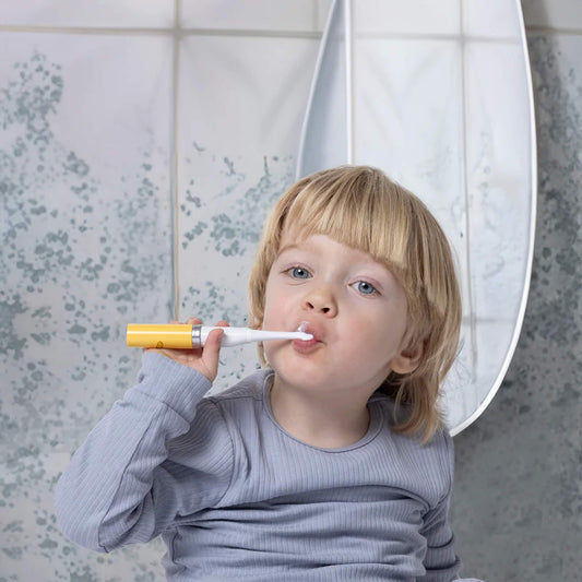 A young child brushing their teeth with a yellow-handled electric toddler toothbrush while standing in front of a bathroom mirror.