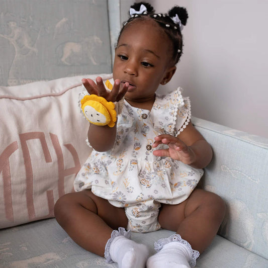A toddler sits on a chair examining the wrist teether on their wrist, lifting their hand to look at the lion-shaped topper.