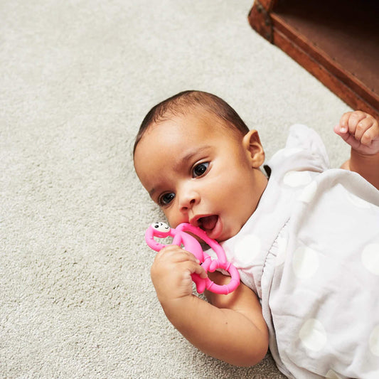 A baby lying on a fluffy cream rug while chewing on a pink monkey-shaped teether held between both hands.