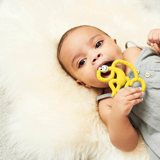 A baby lying on a fluffy cream rug while chewing on a yellow monkey-shaped teether held between both hands.