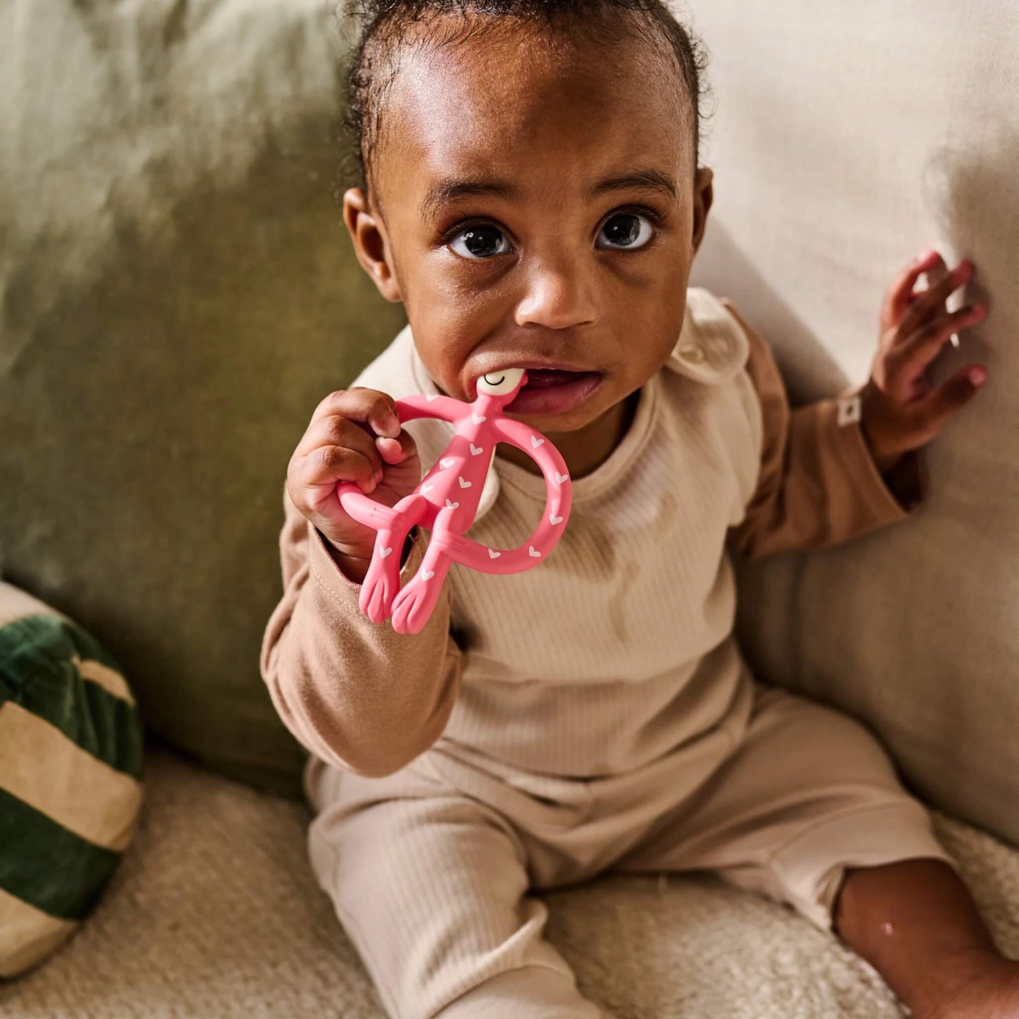 A baby sitting on a sofa holding and chewing a pink heart-patterned silicone teether.