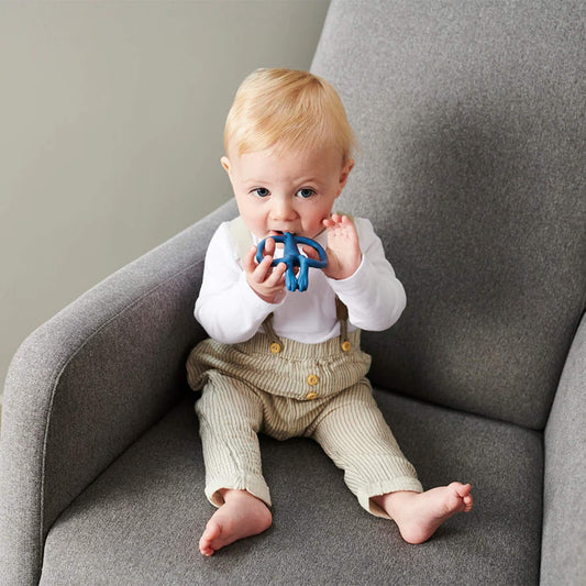 A baby sitting on a grey armchair holding a blue silicone monkey teether and chewing on the textured head.