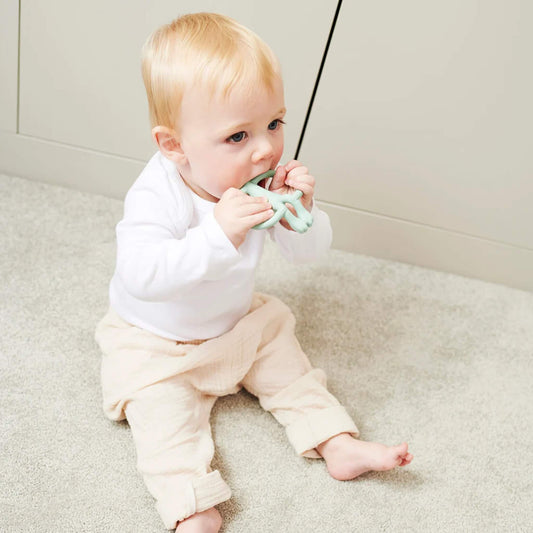 A baby sitting on a light-coloured floor holding a mint green silicone teether with both hands and chewing on the smooth top section. The baby is wearing a white long-sleeved top and pale trousers.