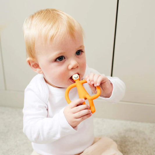A baby sitting on a light-coloured floor holding an orange silicone teether with both hands and chewing on the smooth top section. The baby is wearing a white long-sleeved top and pale trousers.