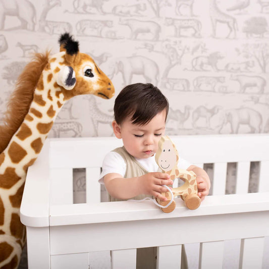 A young child standing at a white cot, holding a wooden giraffe pull toy while a large stuffed giraffe stands beside them.