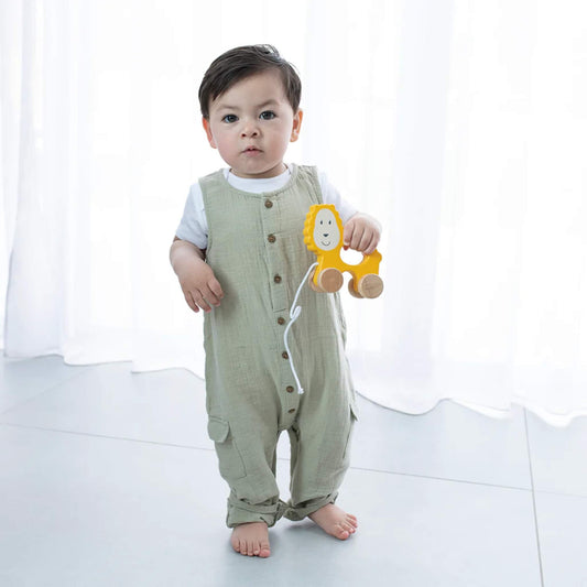 A toddler standing on a light floor holding a small wooden lion pull-along toy by its cut-out centre, with the pull cord hanging loosely.