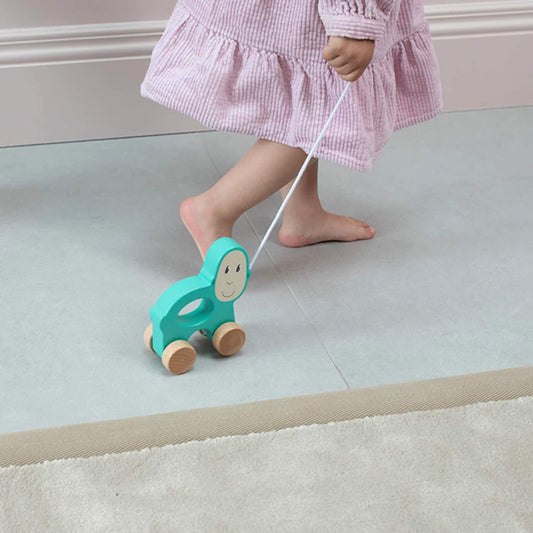 A child walking across a floor pulling the turquoise wooden monkey toy along by its cotton string.