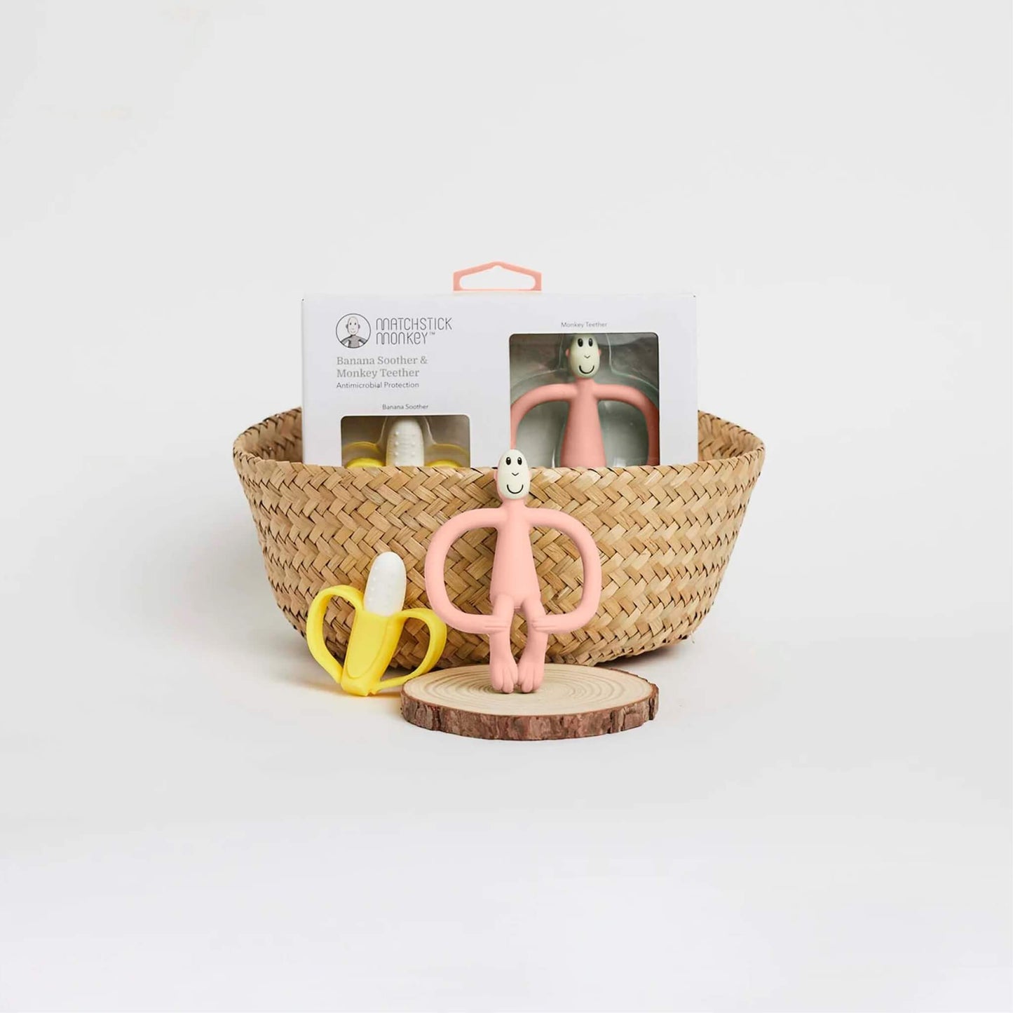 A dusty pink monkey teether and yellow banana soother displayed in front of a woven basket with the boxed set positioned behind them.