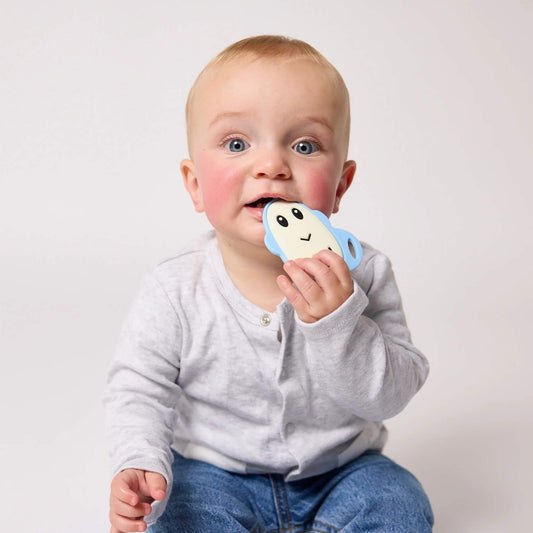 A baby with blue eyes sits wearing a grey top and jeans while holding a flat silicone teether against their mouth.