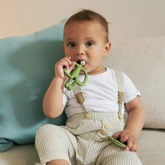 A toddler sitting on a sofa chews an olive animal-shaped silicone teether while holding a matching flat teether.