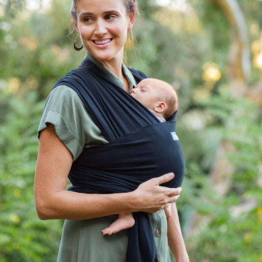 Mother smiling outdoors while carrying her baby close in a black baby wrap carrier.