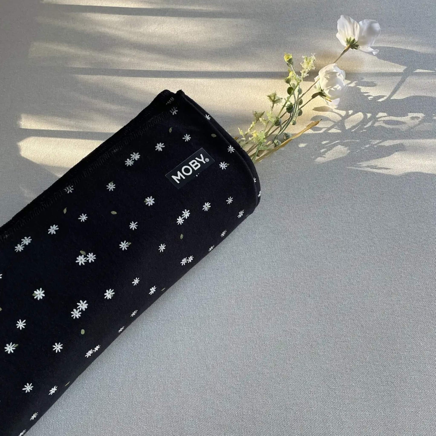 Rolled black baby wrap decorated with tiny white daisies resting on a surface, with small flowers placed beside it in soft natural light.