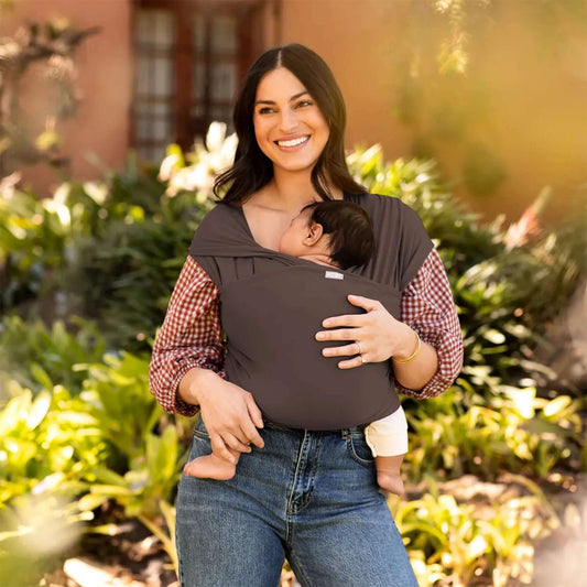 Mother smiling outdoors while carrying her baby in a Hickory brown baby wrap carrier surrounded by greenery.