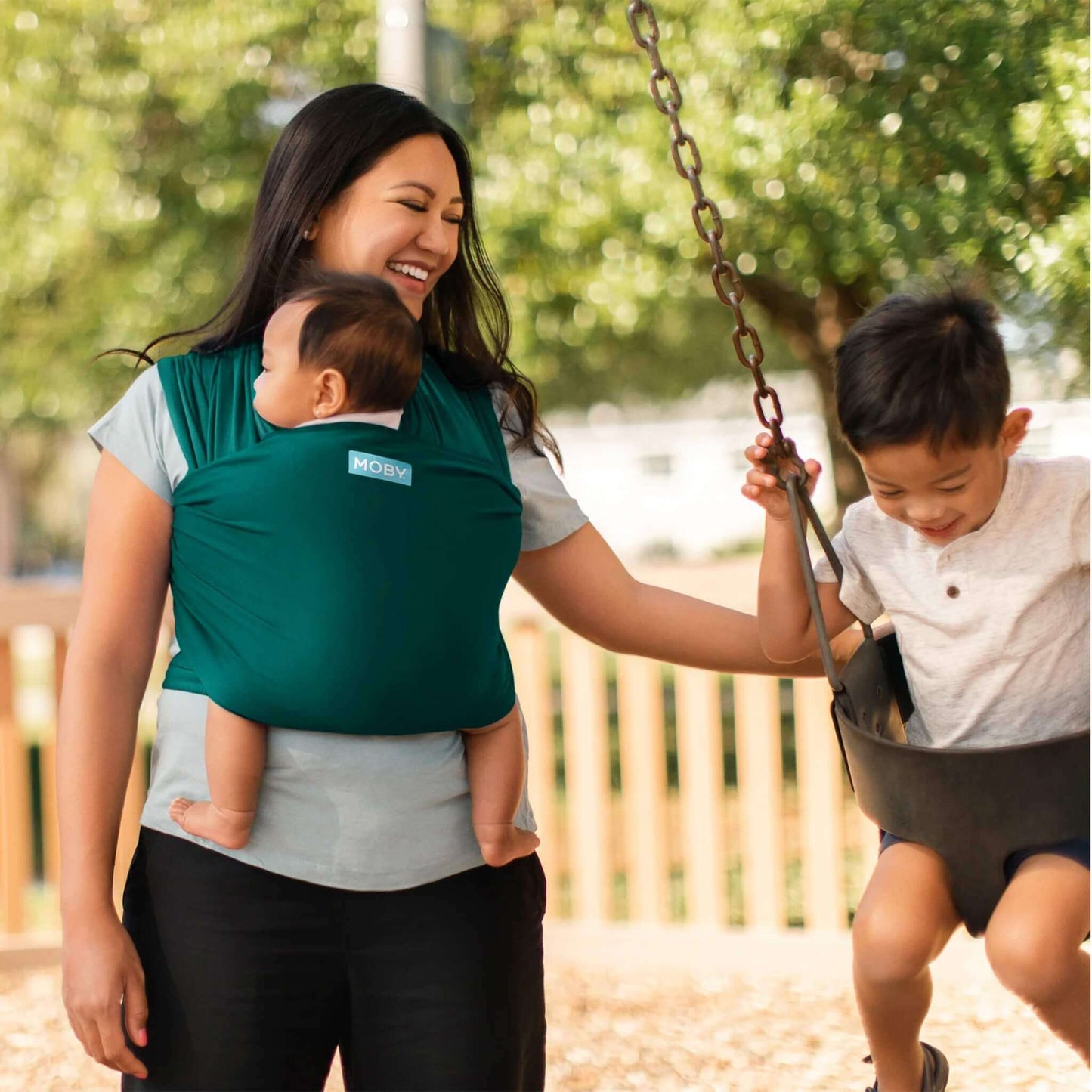 Mother wearing a Pacific green baby wrap carrying her baby while playing with an older child on a swing.