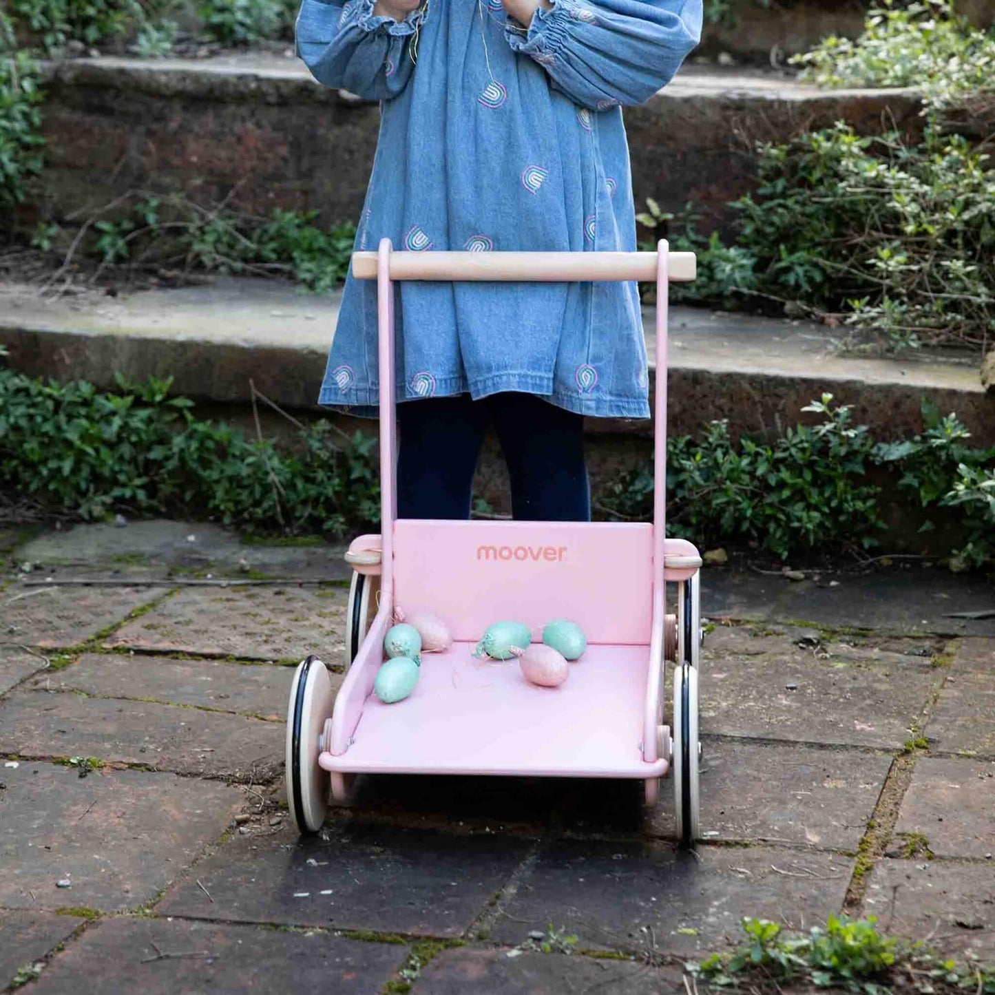 Front view of a pink baby walker filled with pastel toy eggs on a brick patio, with a child standing behind it.
