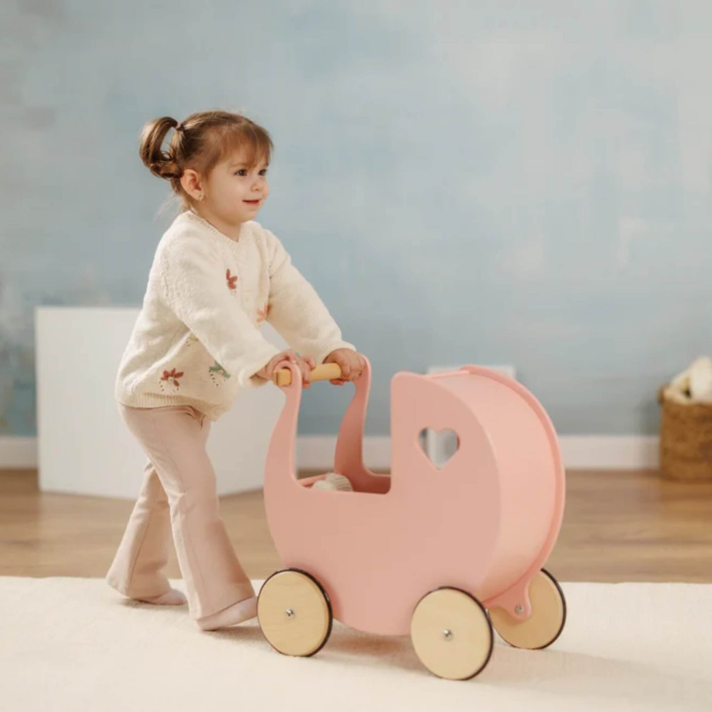 Toddler pushing a pink wooden doll’s pram with a wooden handle across a cream rug in a calm playroom.