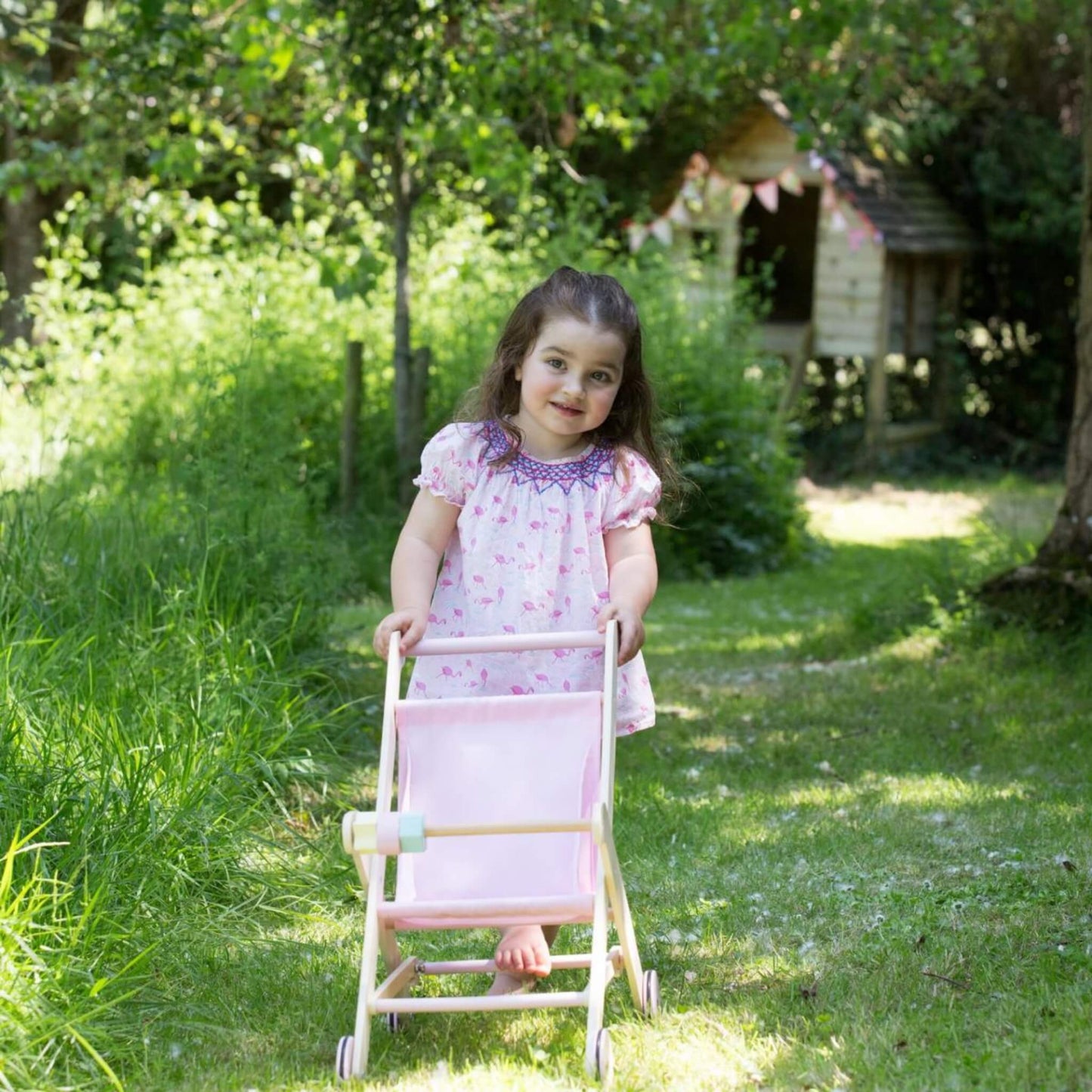 Young girl in a floral dress smiling as she pushes a pink wooden stroller along a garden path.