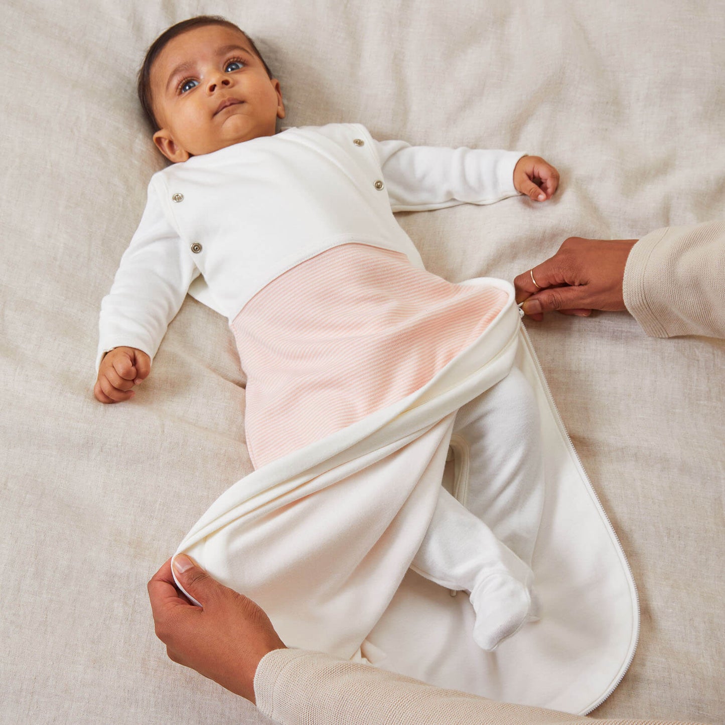 A newborn baby lying on a bed surface while an adult gently opens the side zip of a blush striped swaddle bag to reveal the inner layer.