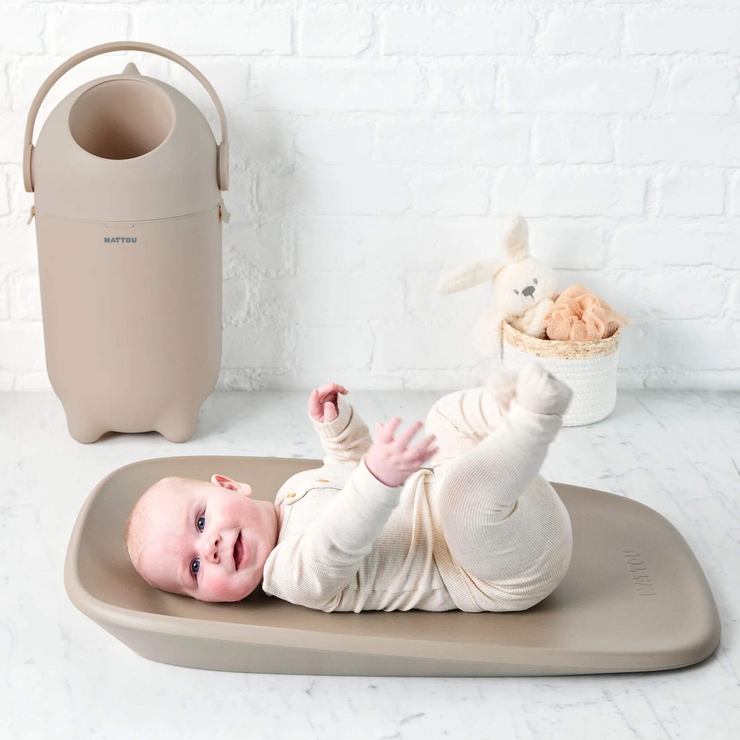 A taupe coloured nappy bin in a nursery setting, placed beside a changing mat with a smiling baby lying on it.