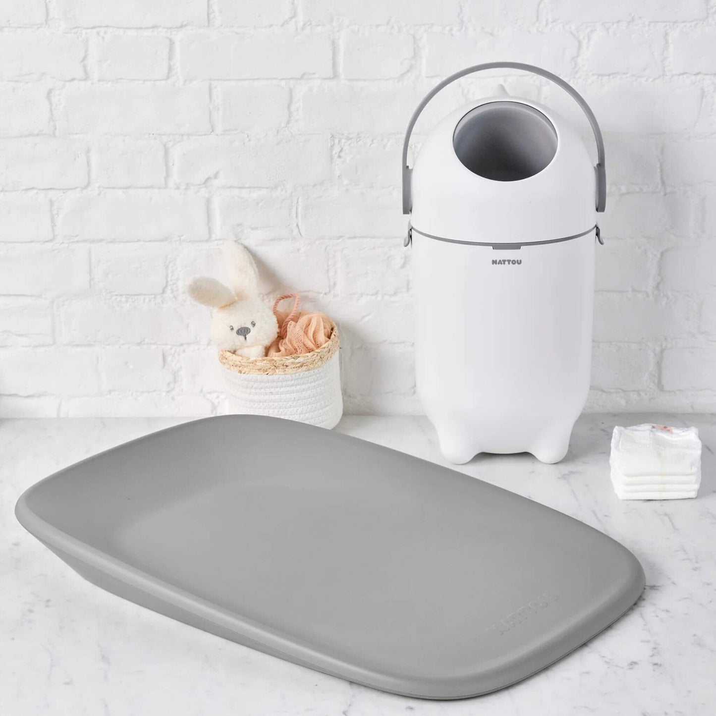 A white nappy bin in a nursery setup, placed beside a grey changing mat and baby essentials.