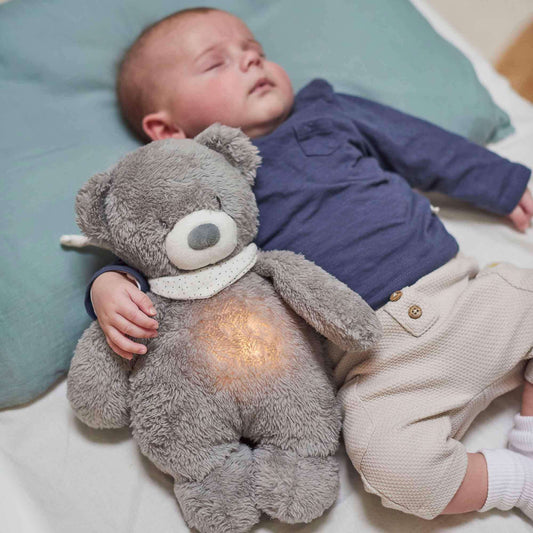 Baby sleeping peacefully while cuddling a plush bear nightlight, which emits a soft warm glow from its belly.
