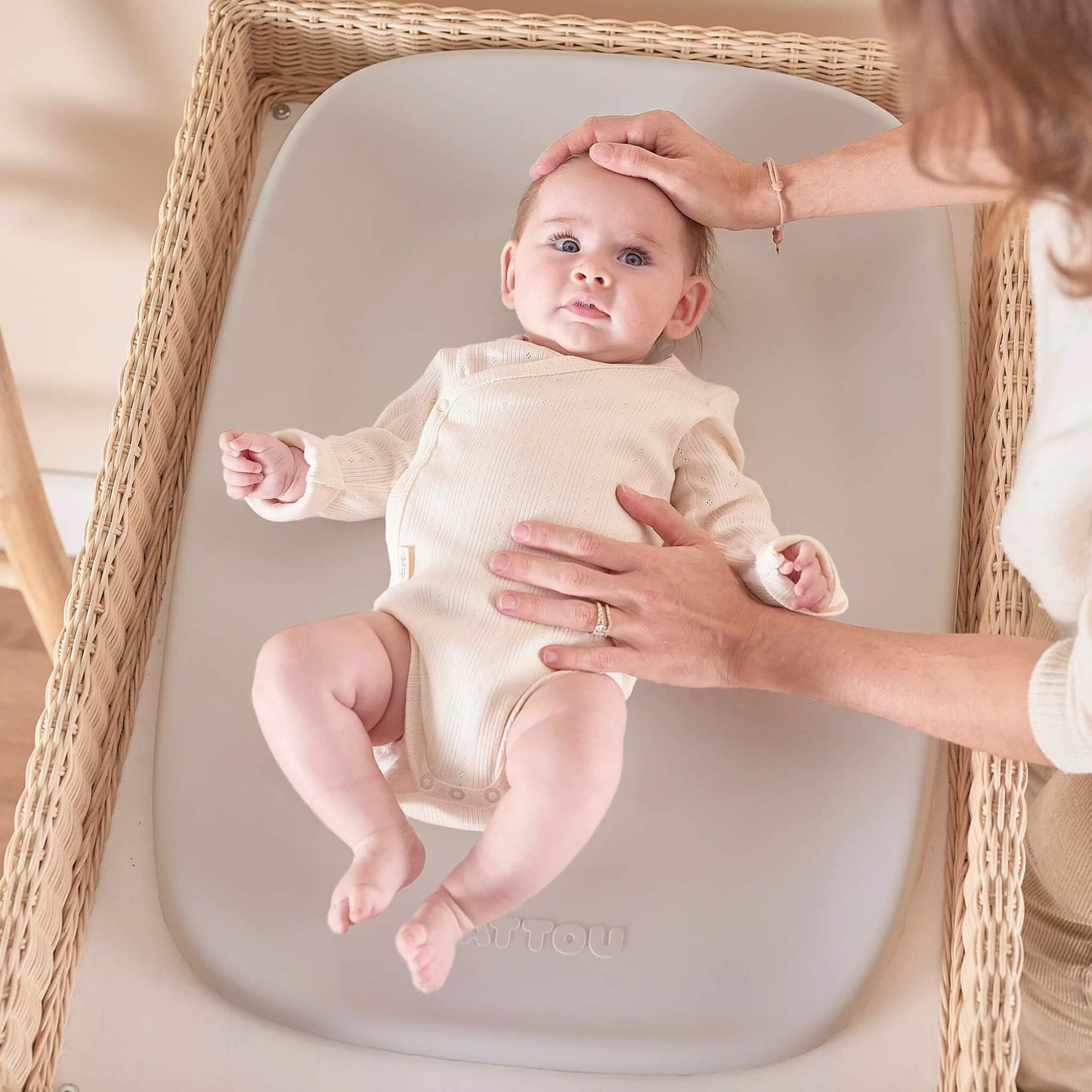 A baby lying on a clay-coloured changing pad placed inside a woven changer basket, with an adult gently supporting the baby’s head and torso.