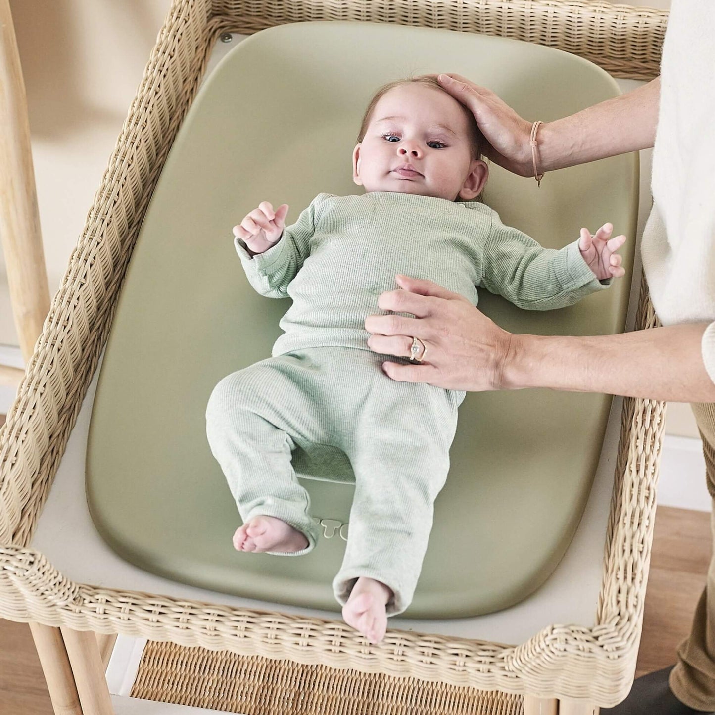 Baby lying on a soft green changing pad while an adult gently supports their head and torso during a nappy change.