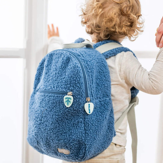 Toddler wearing a soft fabric plush blue teddy Backpack, holding a parent's hand while looking out a window.