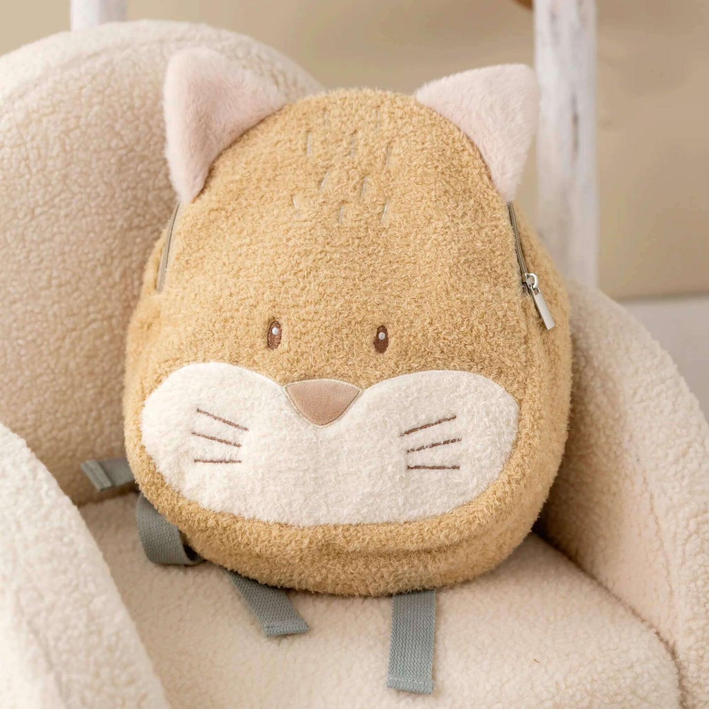 A soft fox-shaped toddler backpack resting on a cream textured chair, showing plush ears, embroidered whiskers and adjustable grey shoulder straps.