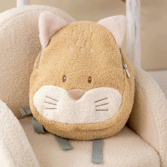 A soft fox-shaped toddler backpack resting on a cream textured chair, showing plush ears, embroidered whiskers and adjustable grey shoulder straps.