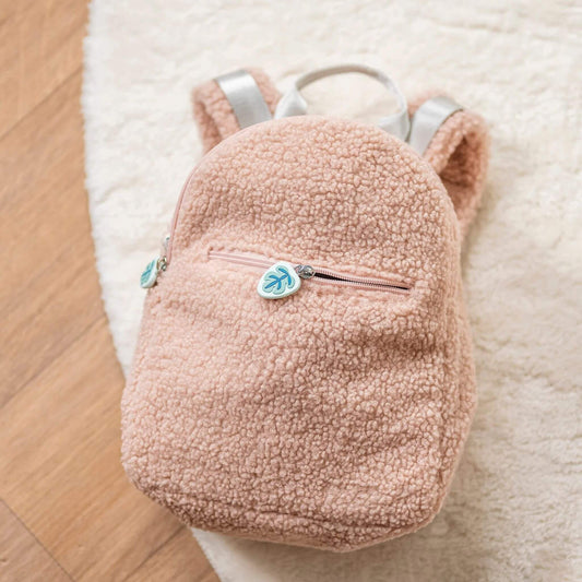 A pink kid's teddy fabric backpack with a soft and plush, textured fabric, zippered front pocket, and adjustable straps, resting on a cosy rug.