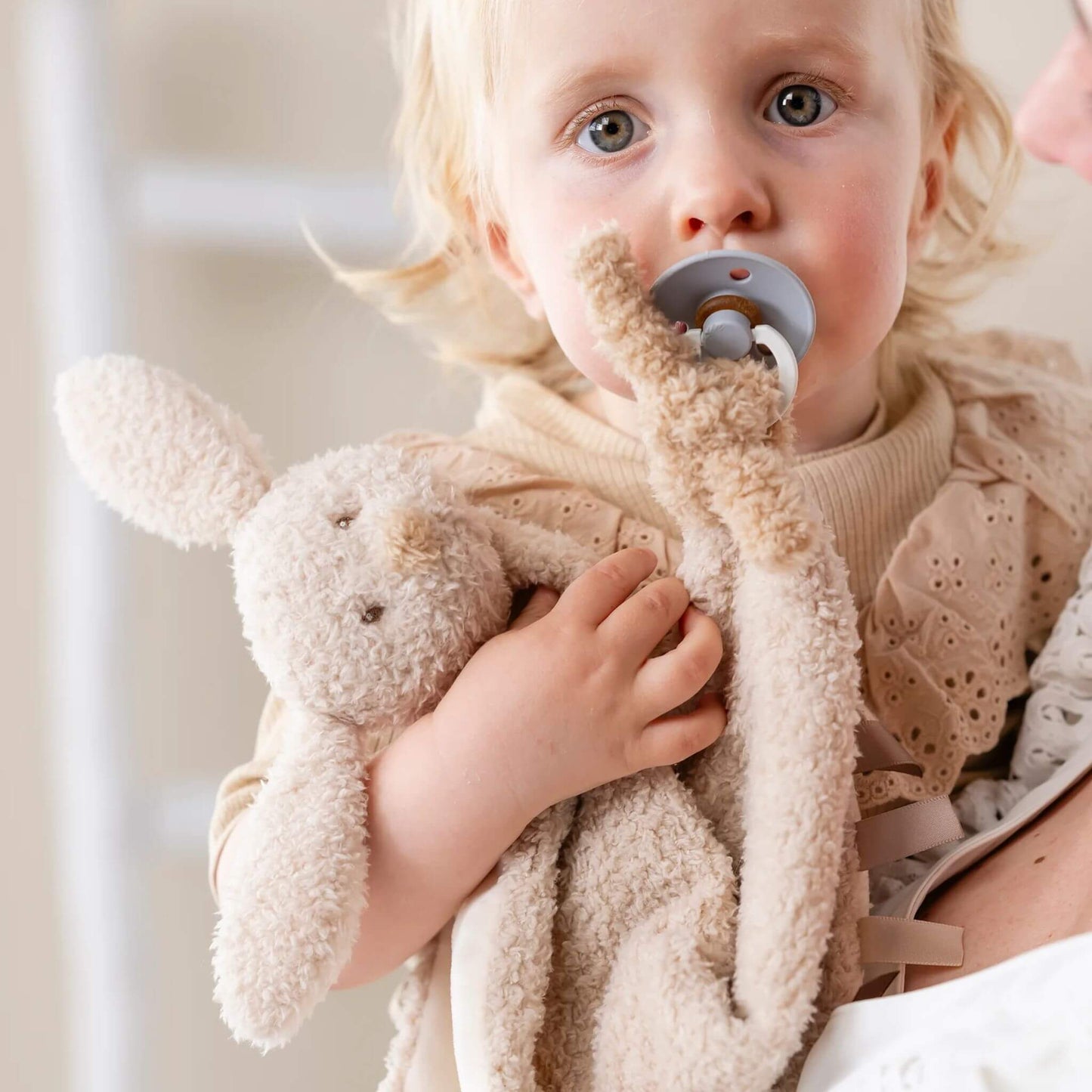 Toddler holding a Bunny design Doudou comfort blanket, showcasing soft beige plush material and comforting design and using the pacifier attachment.