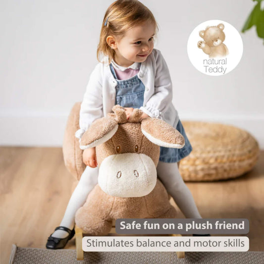 Toddler sitting on a soft beige plush donkey rocker indoors, holding wooden handles while balancing on a curved wooden rocking base.