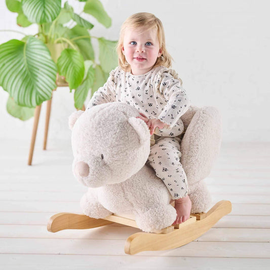 Smiling toddler riding a soft fabric Teddy Bear Rocker in ecru. Set in a bright room with natural wood accents.
