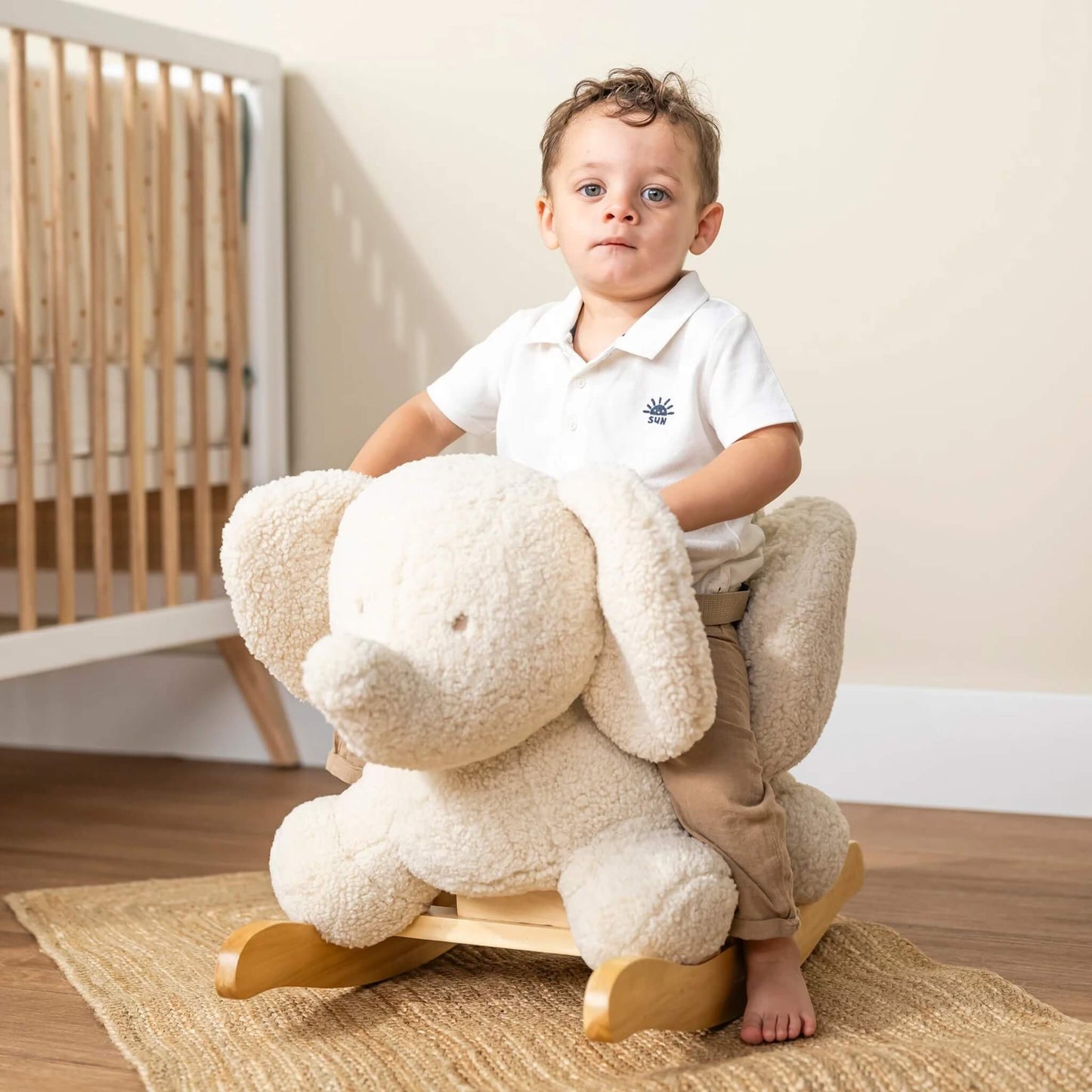 A young child sitting on a plush elephant rocker in a bright nursery, with wooden flooring and a cot in the background.