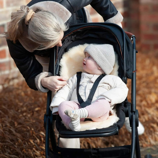 Baby sitting in a pushchair with a sheepskin pram liner, looking up at their mother leaning in beside them outdoors.