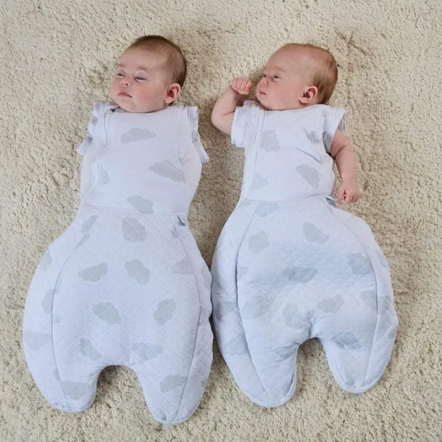 Two babies resting next to each other on a textured rug, wearing cloud-patterned sleep bags, showing one set in arms-in swaddle mode and the other configured with arms out.