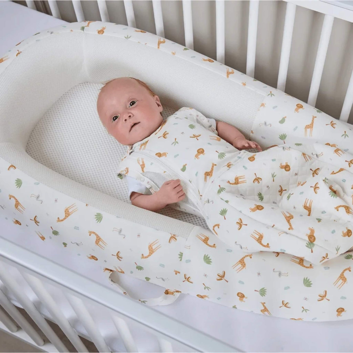 A baby resting in a baby bed while wearing a swaddle to sleep bag, showing a secure swaddle-style top and a shaped lower sleep bag with safari animal print.