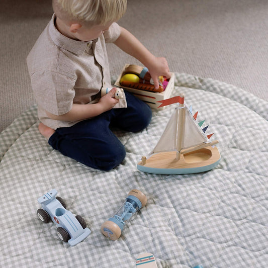 A toddler sitting on a round padded gingham play mat, reaching into a wooden toy crate while holding a small figure, with a toy sailboat nearby.