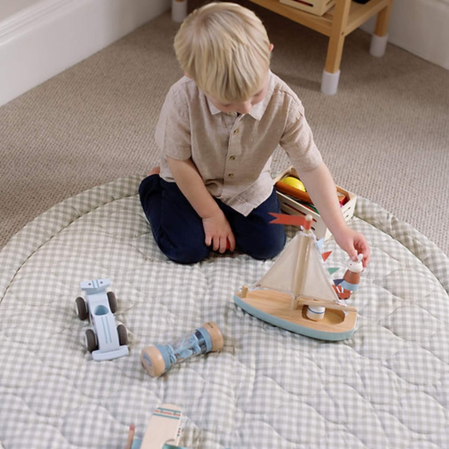 A child kneeling on a padded gingham play mat, placing a small figure into a wooden sailboat toy, with other toys arranged around them.