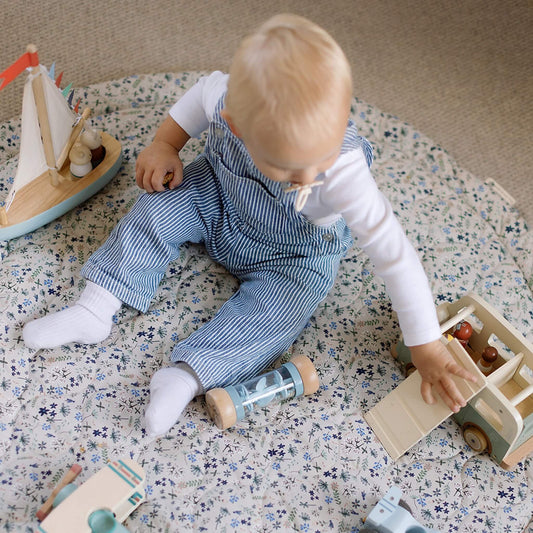 A baby sitting on a padded quilted play mat, reaching for a wooden toy truck, with a rattle and toy car placed nearby on the soft surface.