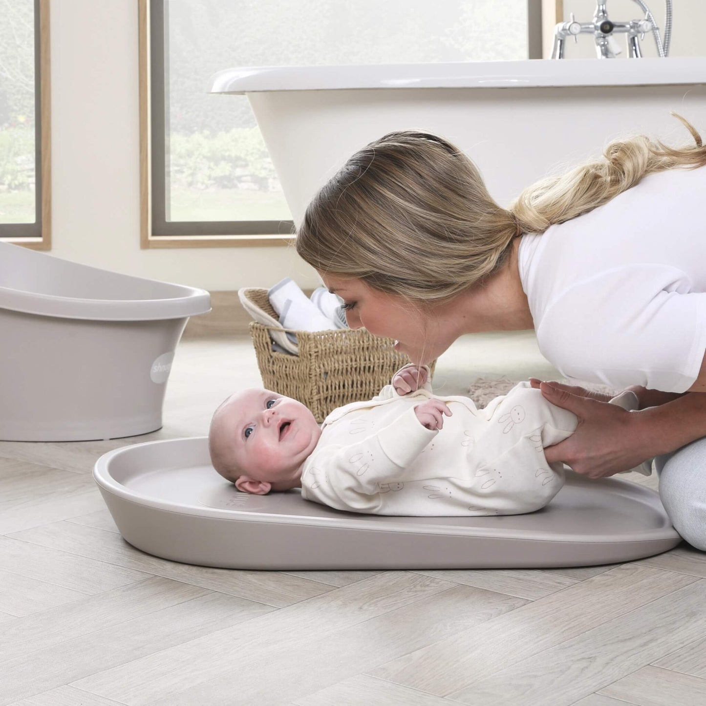 Mother gently wrapping her baby in a hooded towel while the baby lies on a taupe changing mat on a wooden floor.